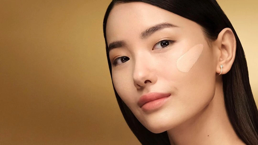 Close-up of a beautiful Asian woman with a flawless complexion, showcasing foundation and makeup against a warm golden background.