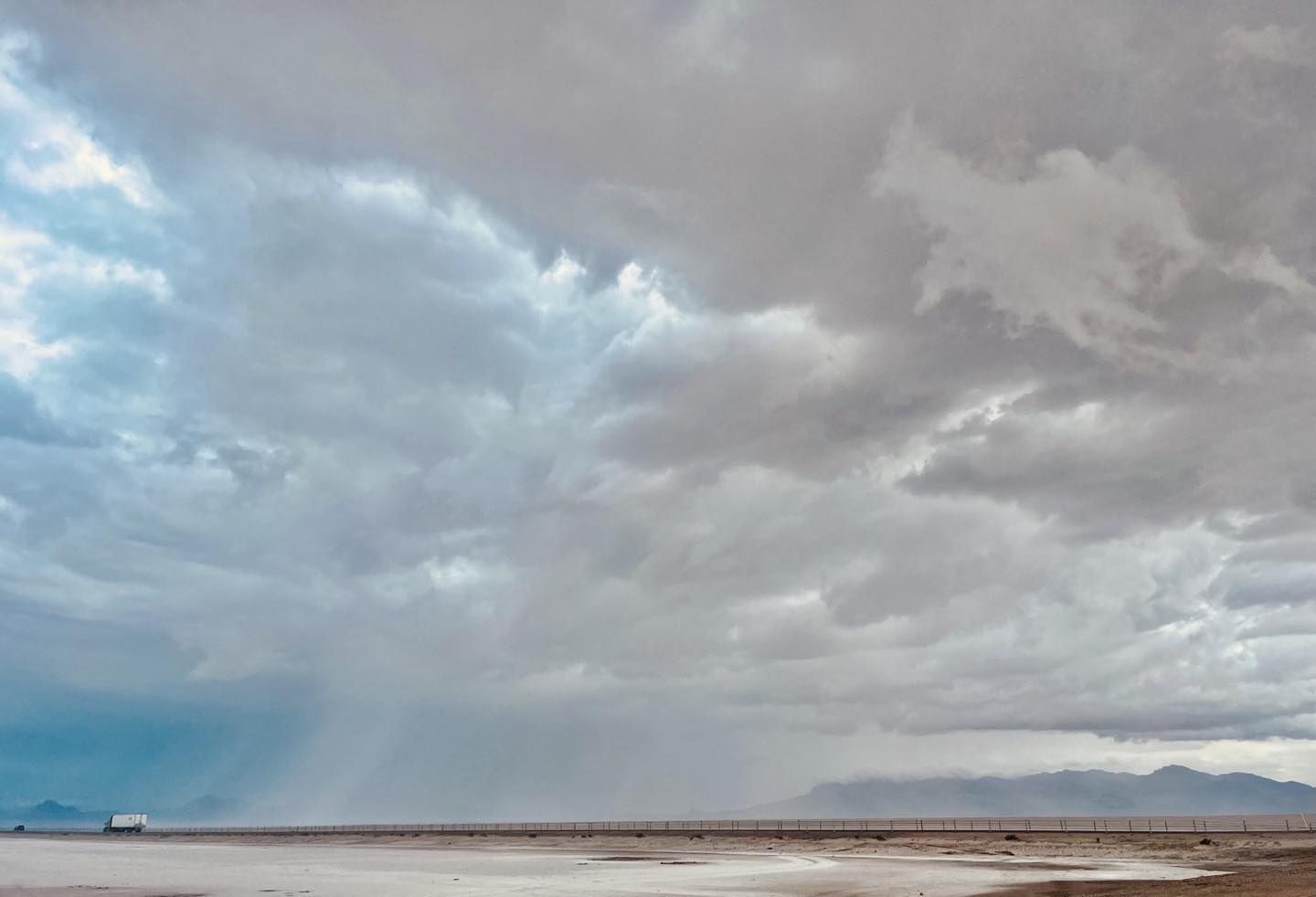 Overcast sky dominates the landscape, with a highway stretching across a flat plain towards distant mountains and a small vehicle.
