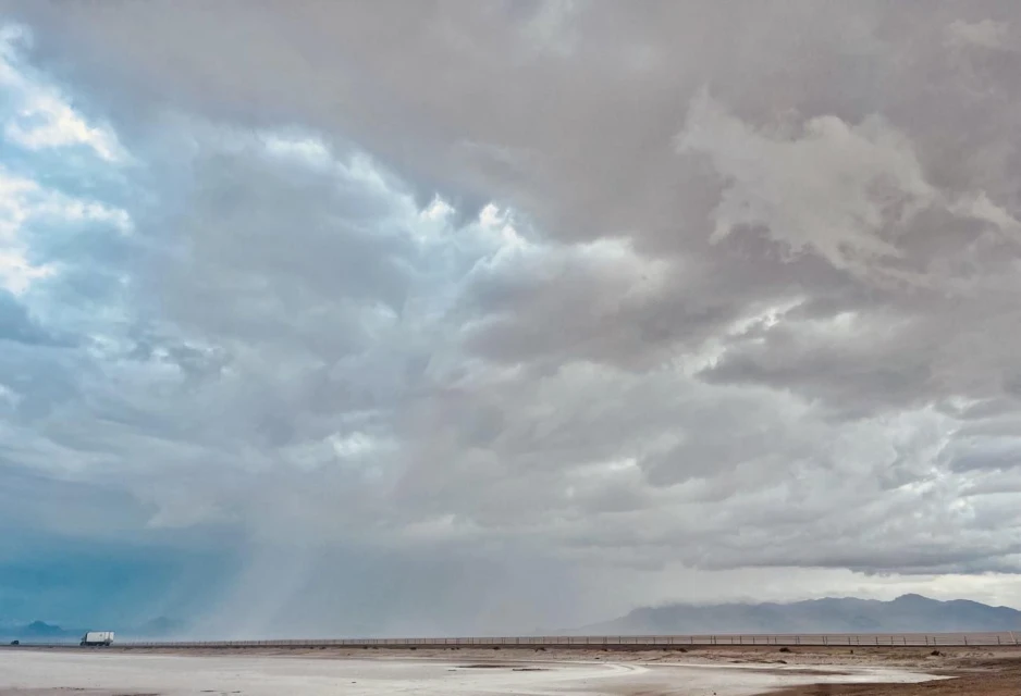 Overcast sky dominates the landscape, with a highway stretching across a flat plain towards distant mountains and a small vehicle.