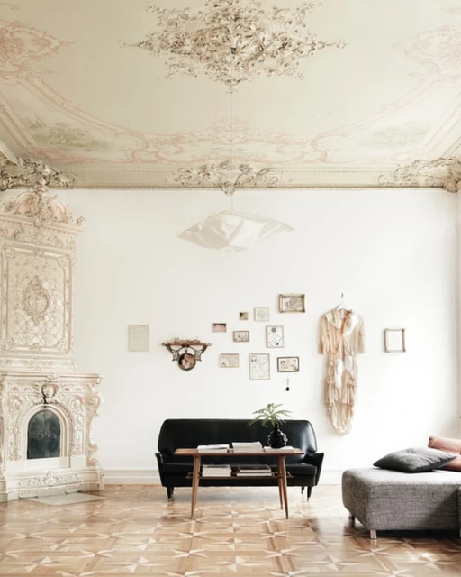 Living room interior with vintage furniture: black sofa, wooden table, stone fireplace, and decorative plaster ceiling in an ornate room.