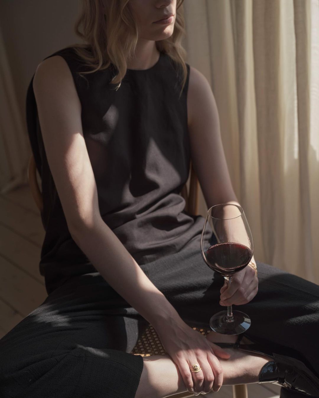 Blonde woman in black sleeveless top and pants sits in soft light holding glass of red wine with curtain background.