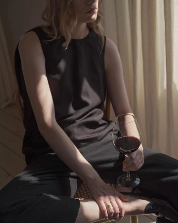 Blonde woman in black sleeveless top and pants sits in soft light holding glass of red wine with curtain background.