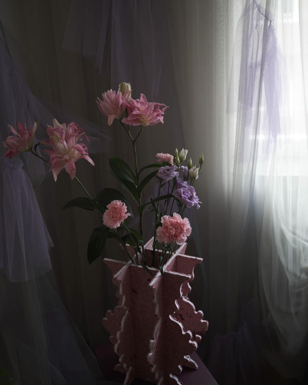 Floral arrangement featuring pink and purple flowers in a pink vase, set against a backdrop of sheer, light-colored curtains.