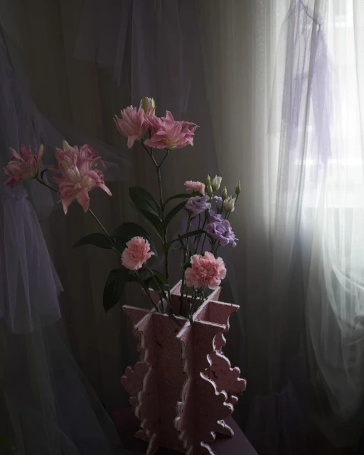 Floral arrangement featuring pink and purple flowers in a pink vase, set against a backdrop of sheer, light-colored curtains.