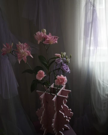 Floral arrangement featuring pink and purple flowers in a pink vase, set against a backdrop of sheer, light-colored curtains.