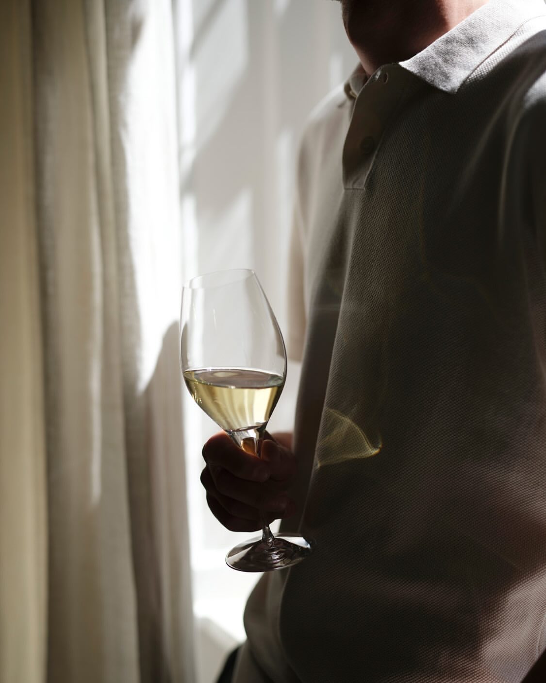 Man indoors holds a glass of white wine; natural light illuminates the scene with sheer white curtains in the background.