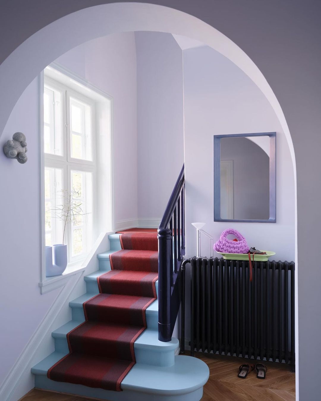 Interior view of a staircase with a red runner, leading up to a window with soft, natural light, and decorative accessories on a radiator shelf.