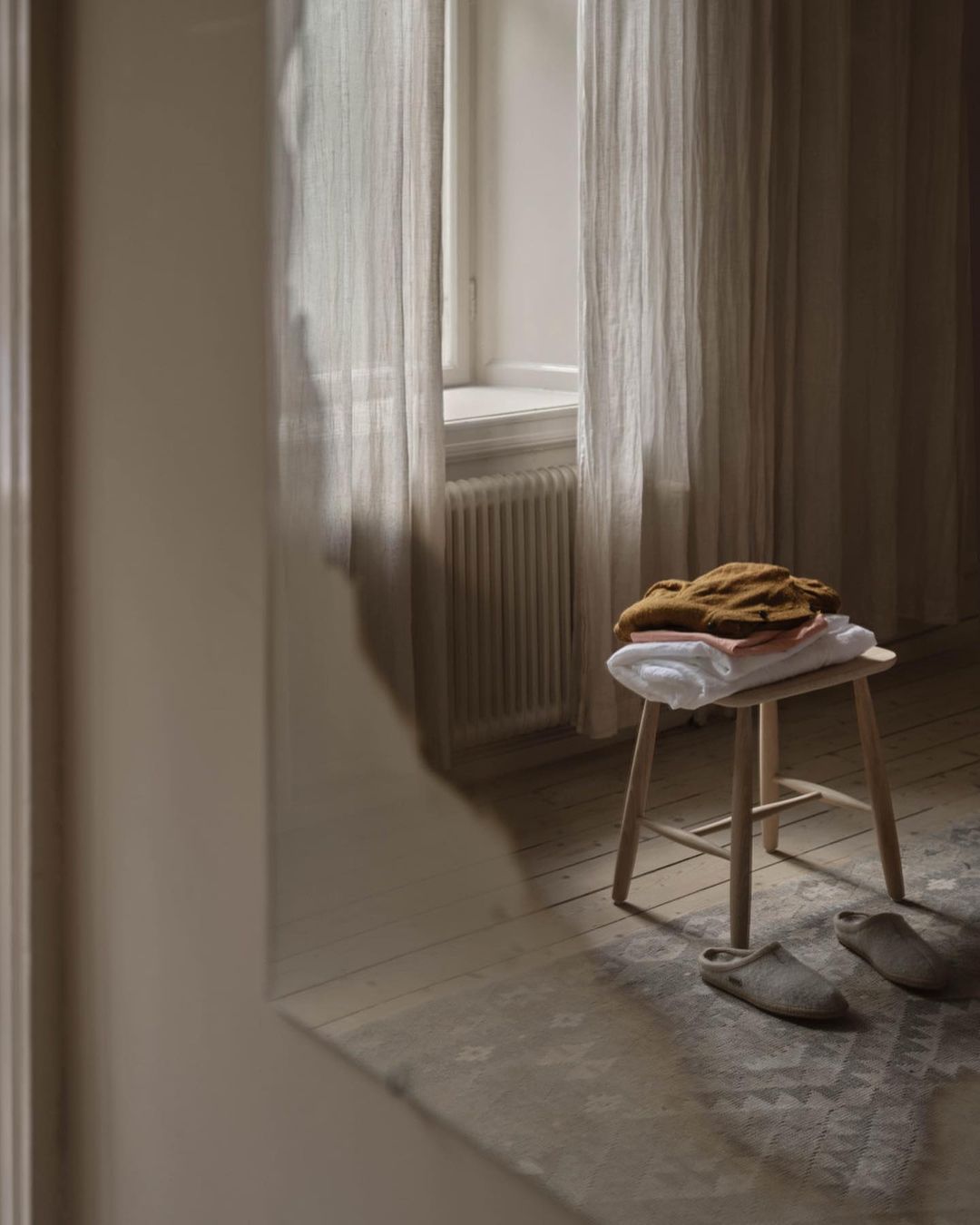 Stack of folded clothes on a stool near a window with sheer curtains, neutral tones, creating a calming and minimalist interior.