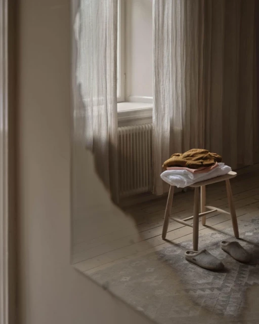 Stack of folded clothes on a stool near a window with sheer curtains, neutral tones, creating a calming and minimalist interior.