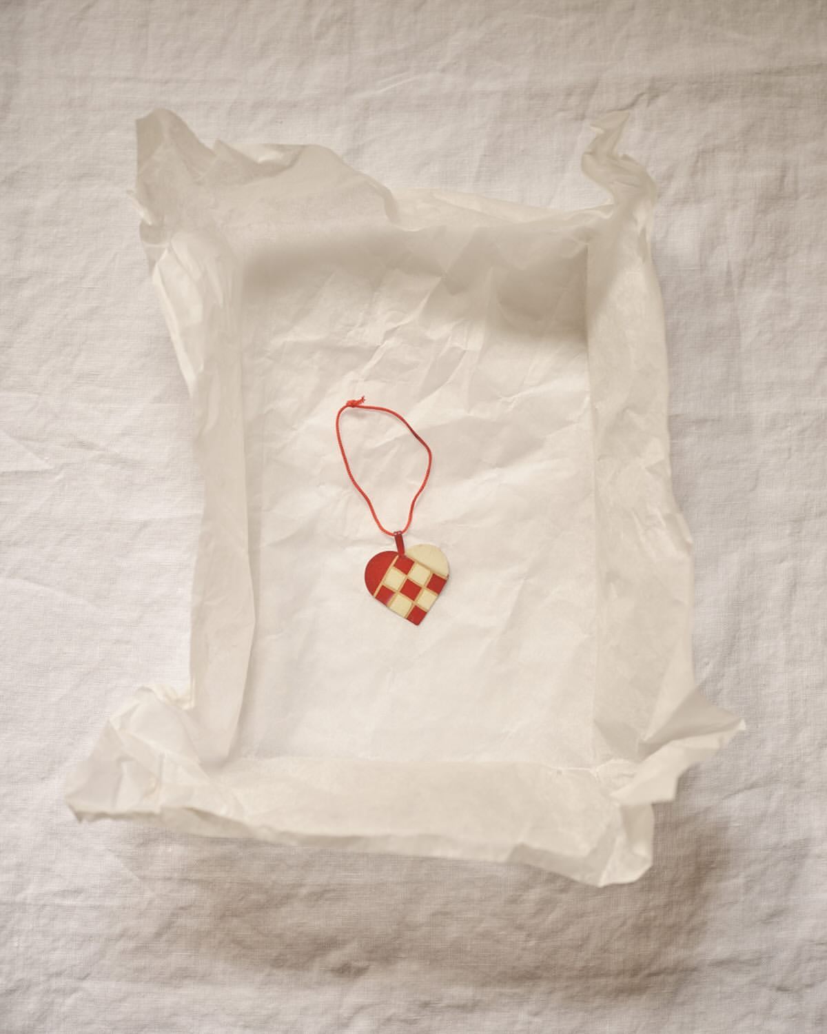 Close-up of a heart-shaped Christmas ornament made of woven red and white paper strips, nestled inside white tissue paper on linen.