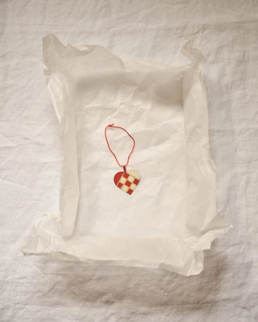 Close-up of a heart-shaped Christmas ornament made of woven red and white paper strips, nestled inside white tissue paper on linen.