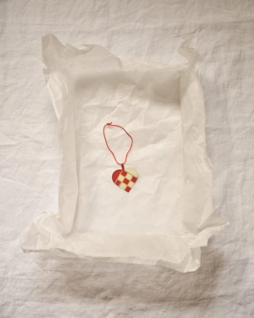 Close-up of a heart-shaped Christmas ornament made of woven red and white paper strips, nestled inside white tissue paper on linen.