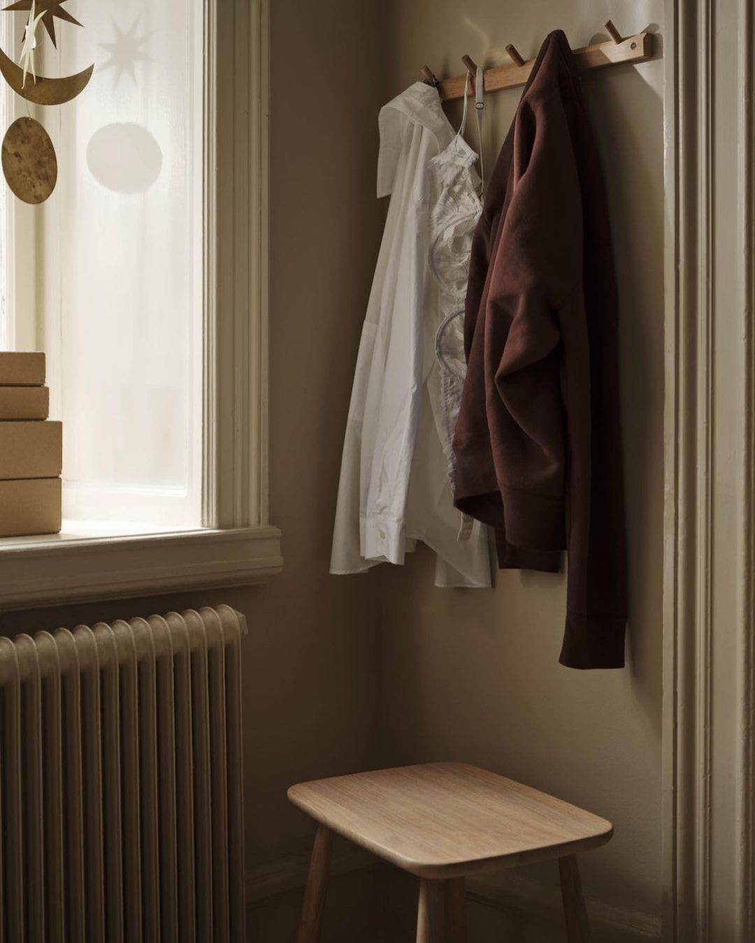 Interior view of a room with a coat rack holding a white shirt and brown jacket, a small wooden stool, and a radiator near a window.