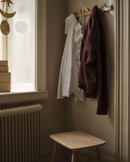 Interior view of a room with a coat rack holding a white shirt and brown jacket, a small wooden stool, and a radiator near a window.