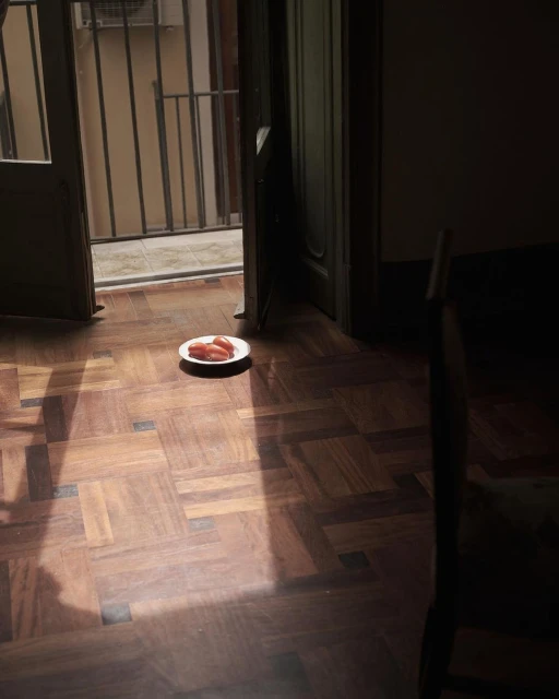 Indoor still life of a plate with three sausages on a parquet floor near an open doorway providing natural light.