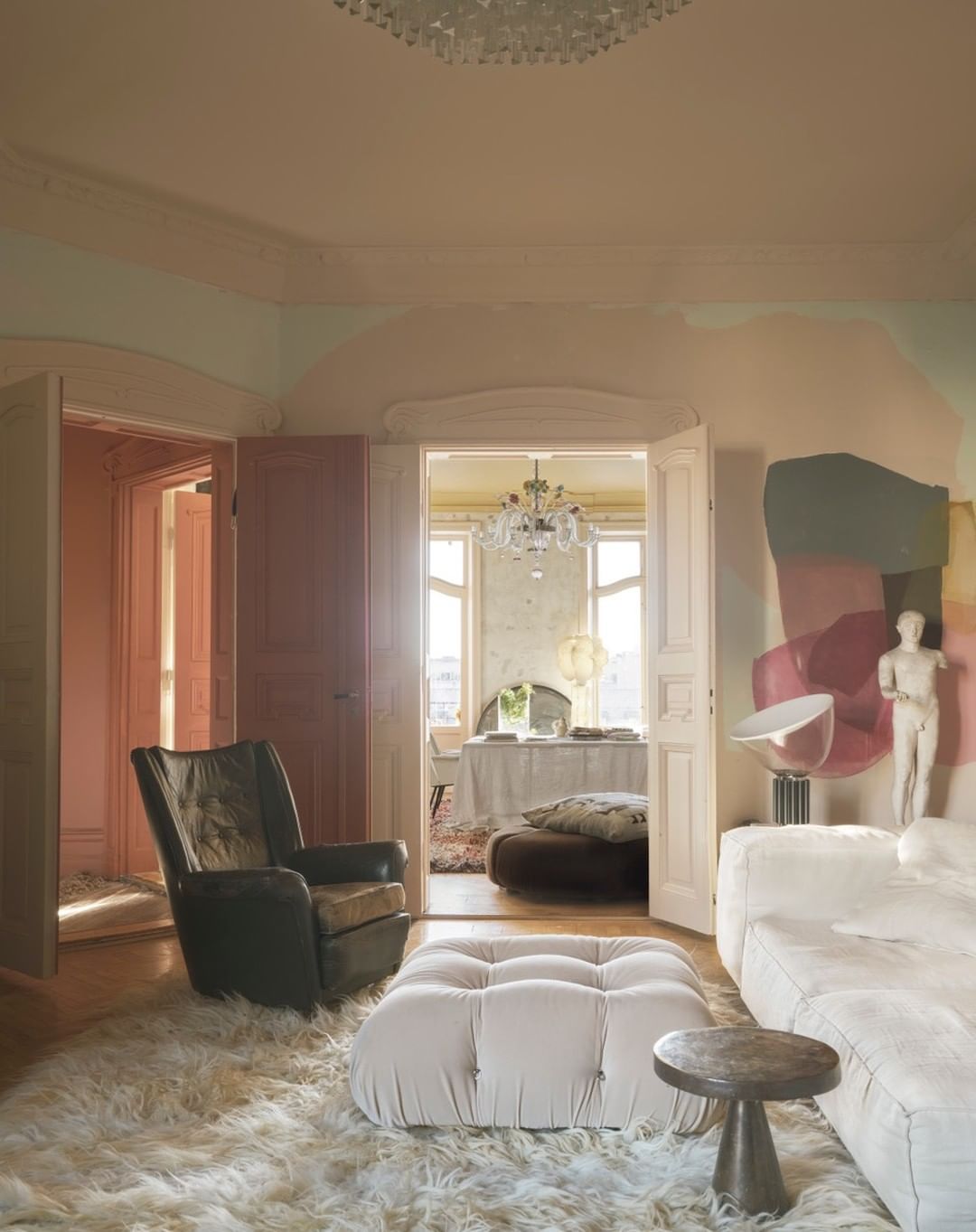 Living room interior with a tan leather chair, fluffy white rug, and tufted ottoman near a round side table in front of a white sofa.