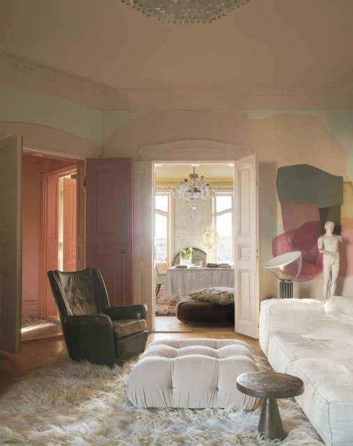 Living room interior with a tan leather chair, fluffy white rug, and tufted ottoman near a round side table in front of a white sofa.