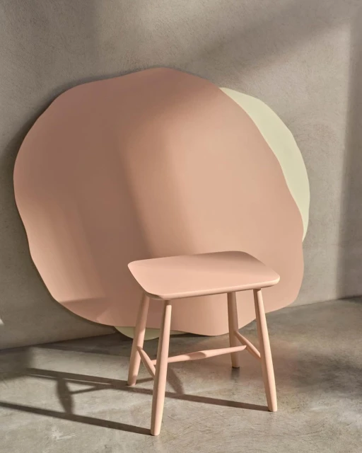 A pink stool sits in front of a pink and beige abstract wall decoration, casting a shadow on a grey floor and wall in a studio shot.