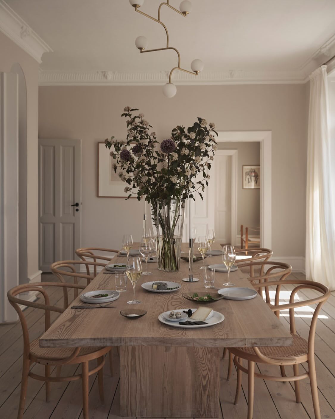 Dining room with wooden table, chairs, vase of flowers, plates, glasses, and a modern chandelier in a bright, neutral-toned home interior.
