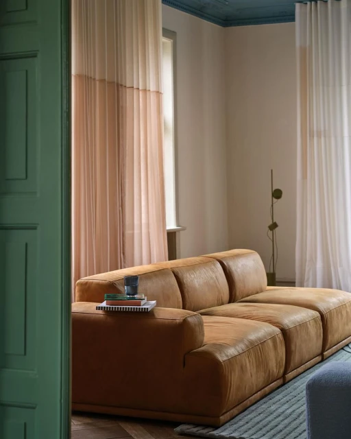 Living room interior featuring a brown leather couch, window with beige curtains, and modern design elements against neutral wall.