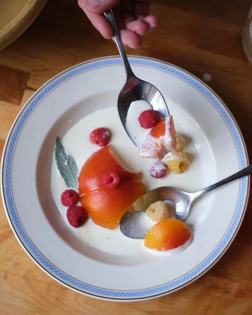 Overhead shot of a plate with persimmon and raspberries covered with cream, with a hand holding a spoonful of the dessert.