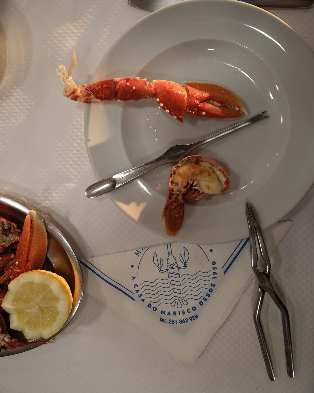 Overhead view of a white plate containing cooked lobster, accompanied by a fork, on a table with a patterned napkin and lemon.