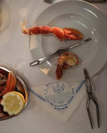 Overhead view of a white plate containing cooked lobster, accompanied by a fork, on a table with a patterned napkin and lemon.