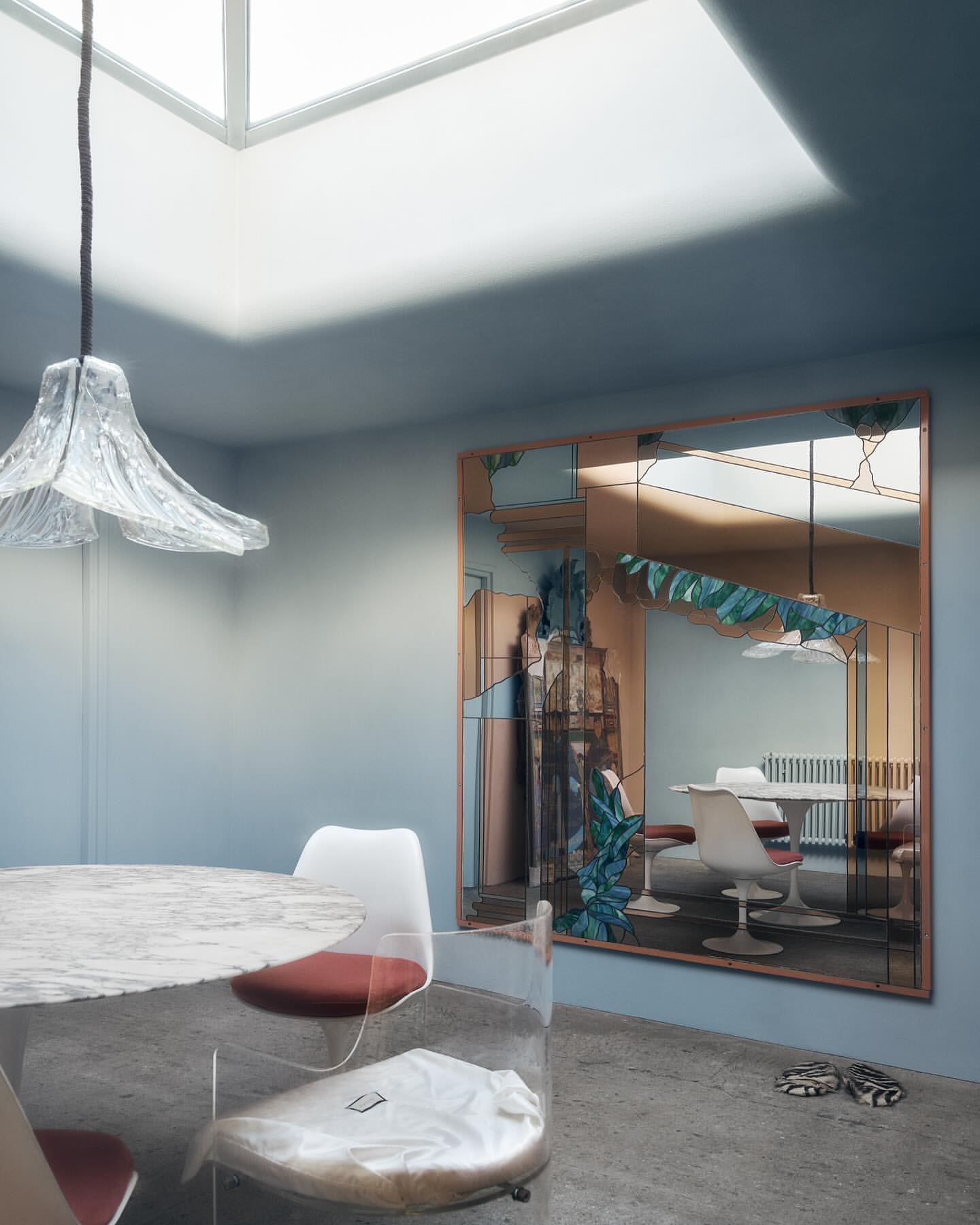 Interior with a round table and clear chairs, a decorative mirror reflecting another table and chairs, and a skylight overhead.