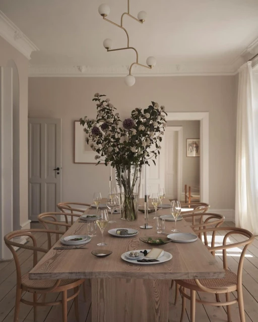Dining room featuring a long wooden table set for a meal with plates, glassware, flowers in a vase, and bentwood chairs.