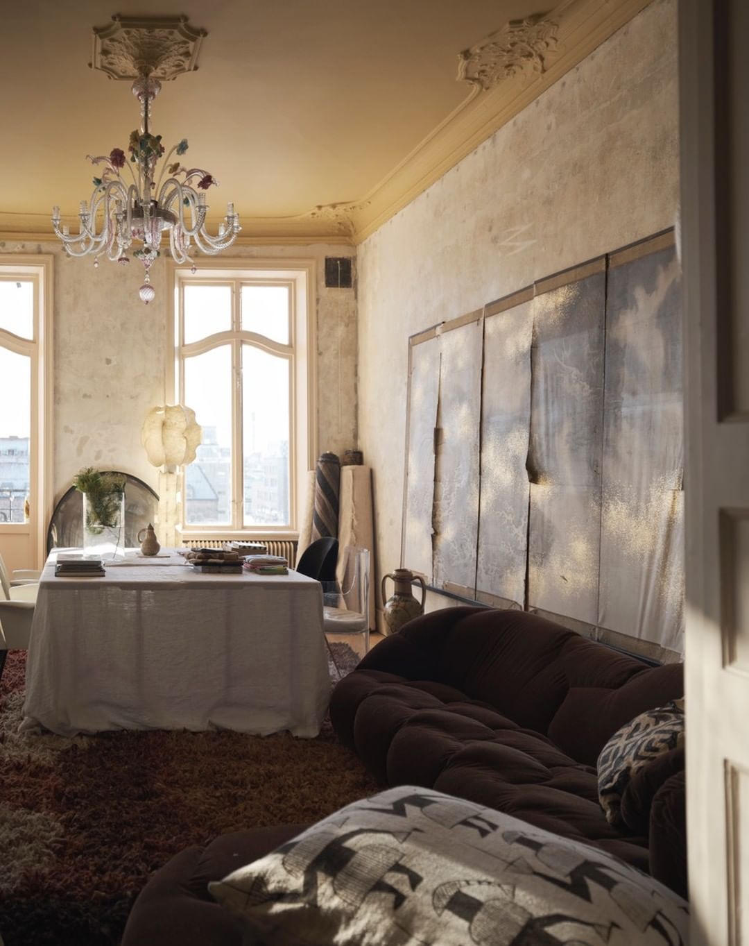 Interior of a room featuring a brown couch, table with white tablecloth, chandelier, decorative vases, and large windows.