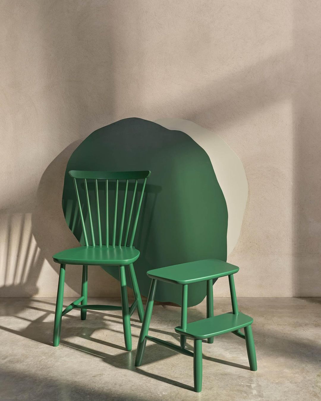 A green Windsor chair and step stool are set against a tan textured wall with green and cream accents, illuminated by soft, indirect light.