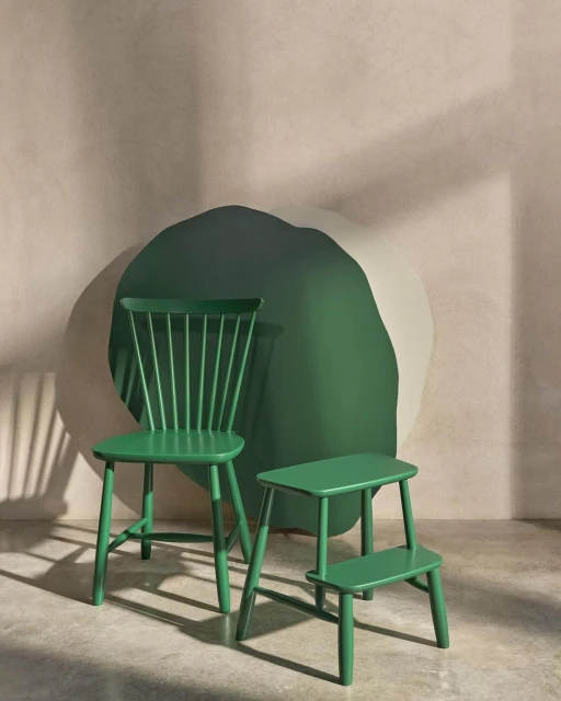 A green Windsor chair and step stool are set against a tan textured wall with green and cream accents, illuminated by soft, indirect light.
