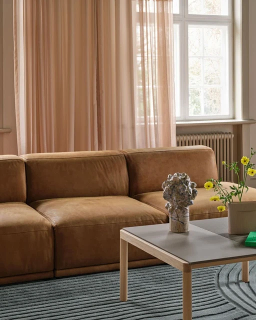 Living room interior featuring a brown leather sofa, a grey coffee table with a vase, and neutral curtains in front of a window.