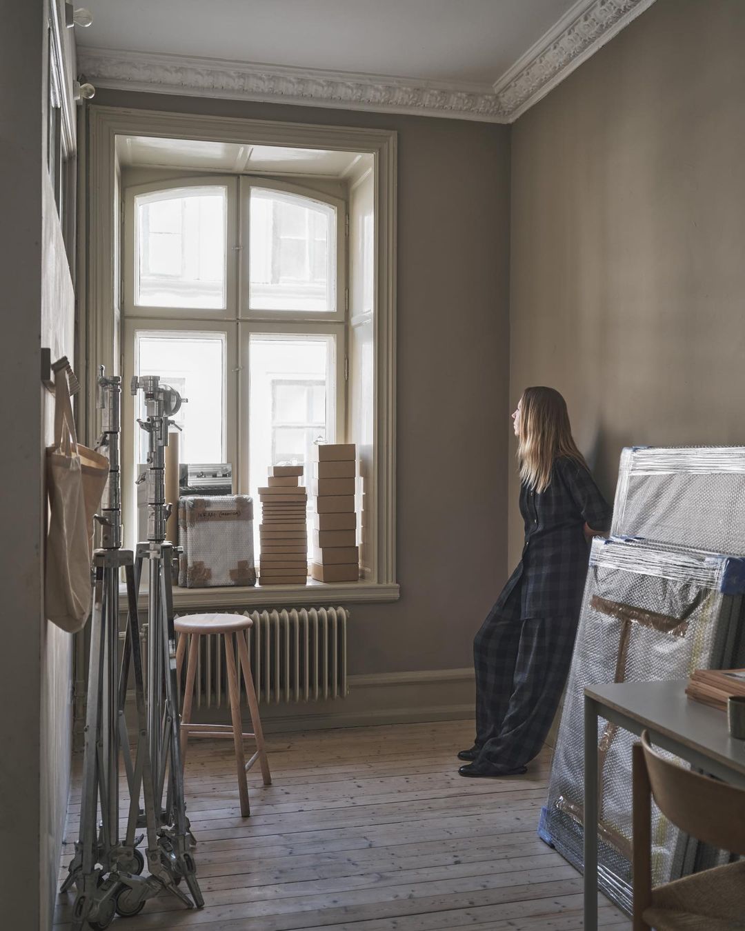 Blonde woman in patterned pantsuit leans against wall in room with lighting equipment, boxes, stool, radiator, and large bright window.