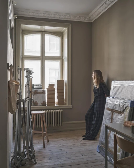 Blonde woman in patterned pantsuit leans against wall in room with lighting equipment, boxes, stool, radiator, and large bright window.