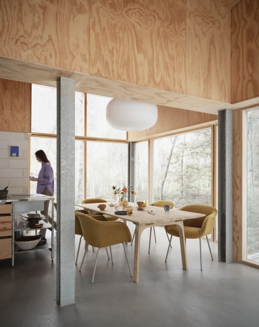 Dining area with a light wood table surrounded by yellow chairs, a woman in the background, and natural light from large windows.