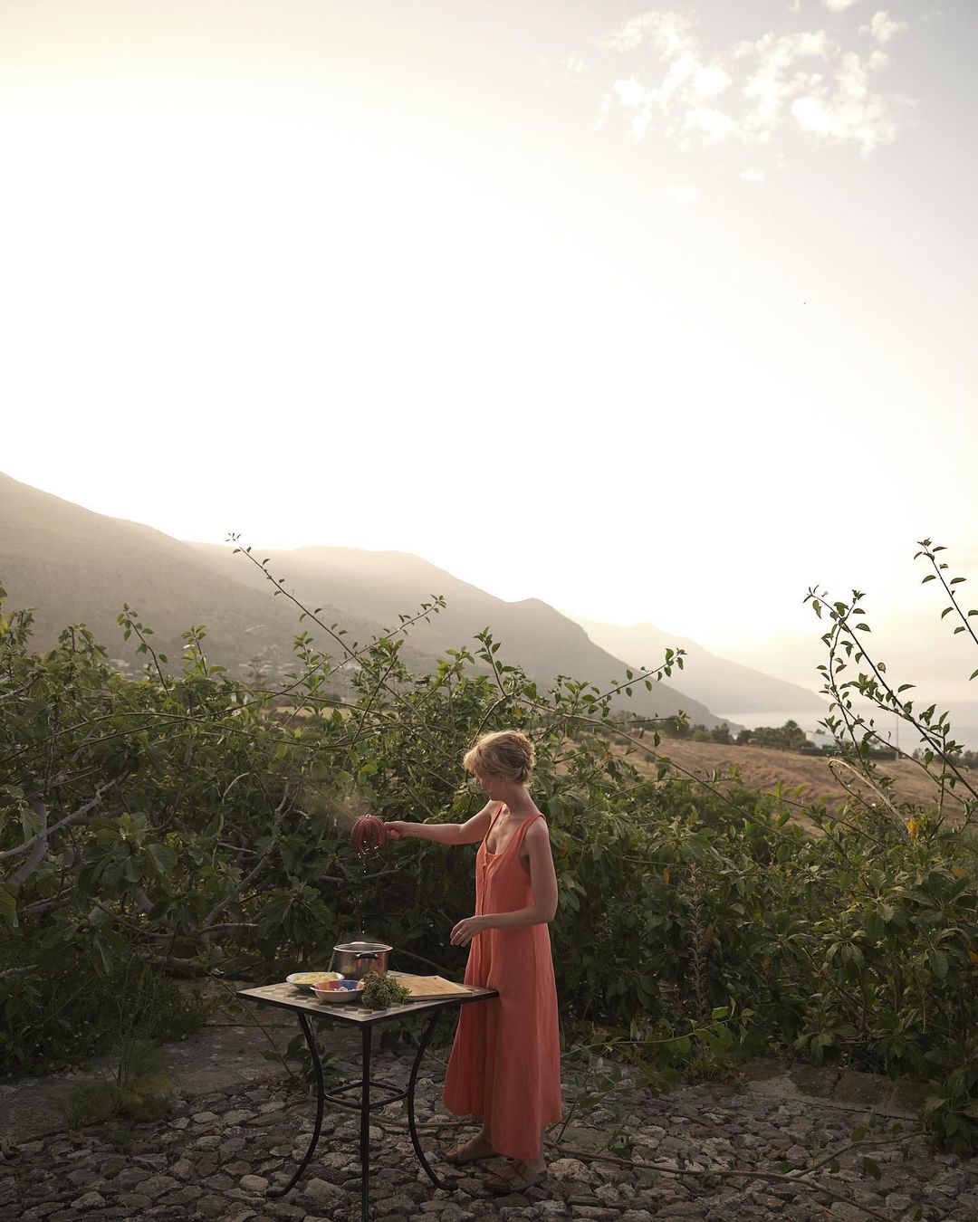 Woman in orange dress stands at table with food, preparing meal outdoors with mountains in background at sunset.