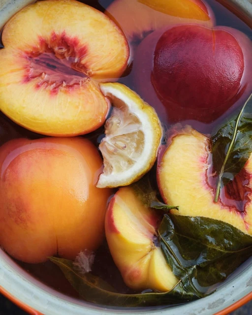 Overhead shot of sliced peaches and lemon in a bowl of liquid, with dark green leaves, showcasing a vibrant and fresh food presentation.