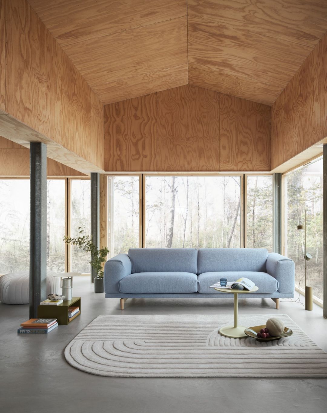 Minimalist living room interior with a light blue sofa, round patterned rug, and plywood walls, flooded with natural light.