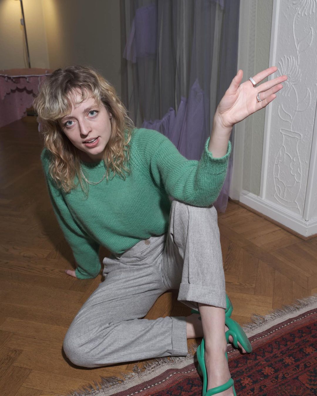 Indoor portrait of a young adult woman with curly blonde hair, wearing a green sweater, gray pants, and green shoes, sitting on a wood floor.