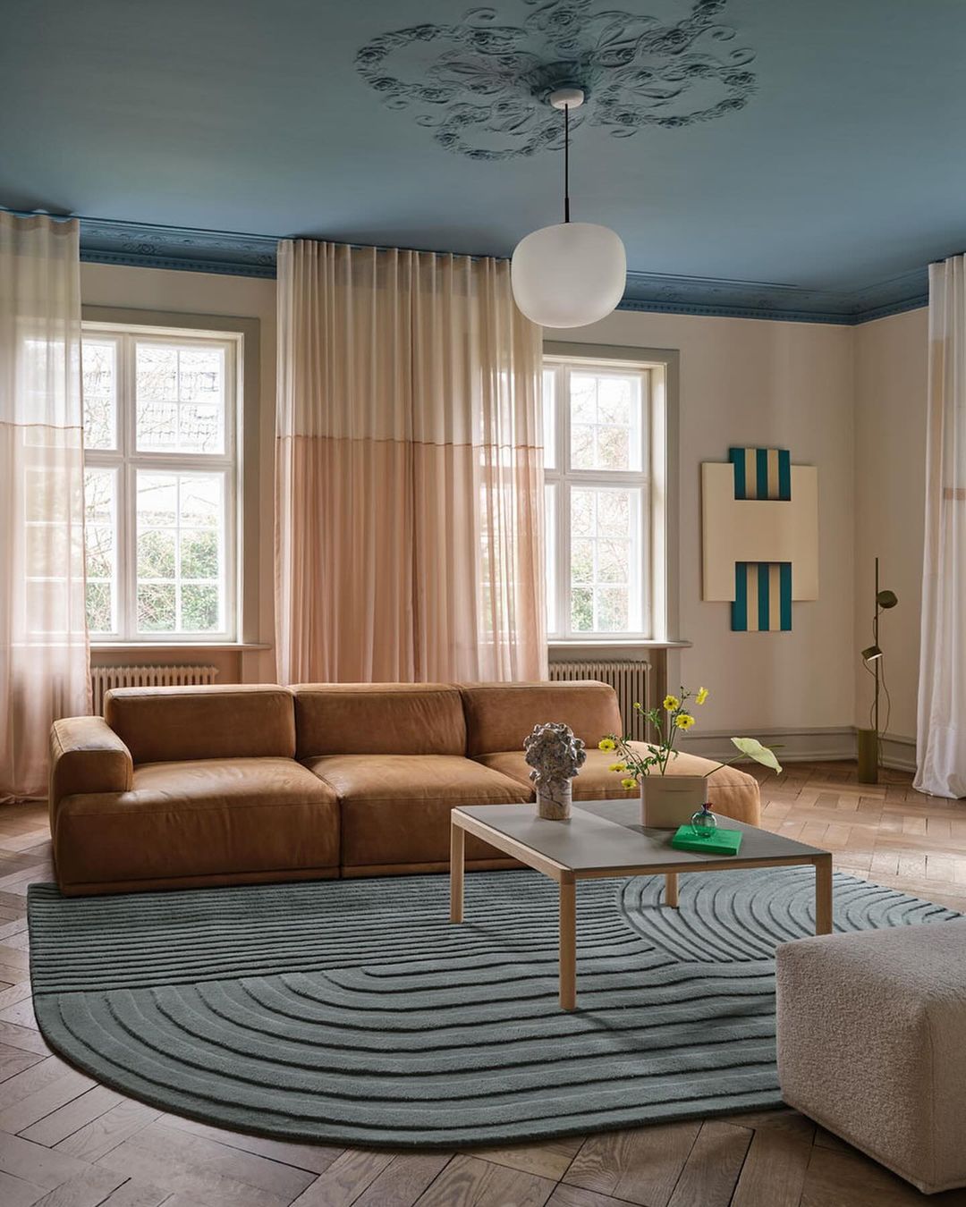Living room interior featuring a brown sectional sofa on a blue-and-gray rug, a concrete coffee table, sheer curtains, and hardwood floors.