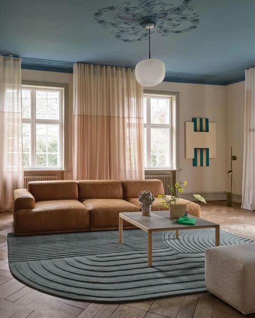 Living room interior featuring a brown sectional sofa on a blue-and-gray rug, a concrete coffee table, sheer curtains, and hardwood floors.