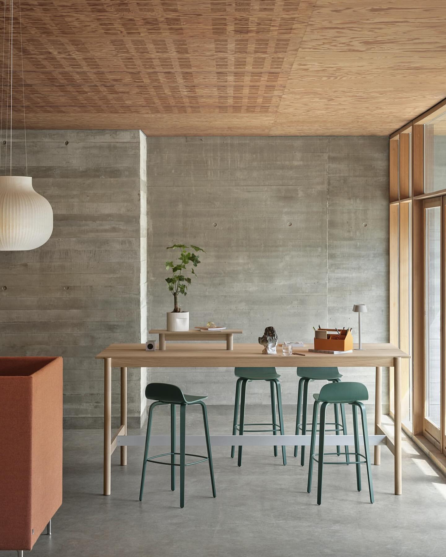 Interior design featuring a light wood table with green stools, concrete wall, wood ceiling, modern lighting, and a minimalist aesthetic.