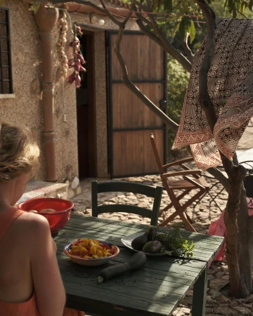Woman preparing food at outdoor table, featuring tomatoes, zucchini, herbs, and a rustic stone house with hanging garlic.