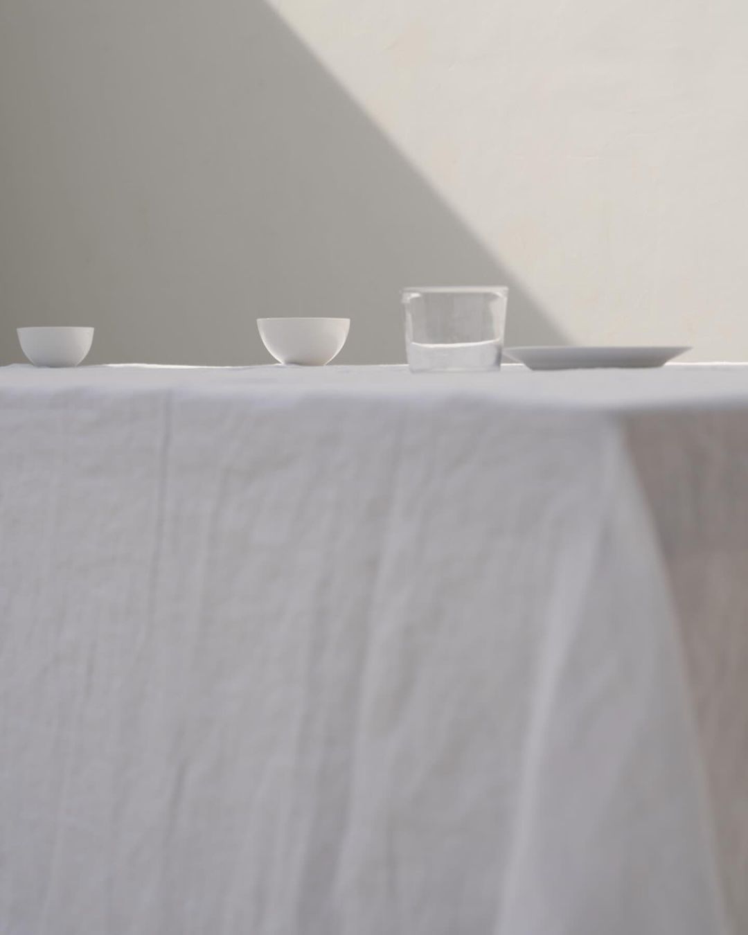 Minimalist still life: White bowls, a glass of water, and a plate on a white tablecloth, bathed in soft natural light against a neutral wall.