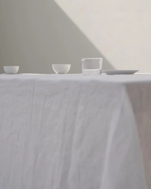 Minimalist still life: White bowls, a glass of water, and a plate on a white tablecloth, bathed in soft natural light against a neutral wall.