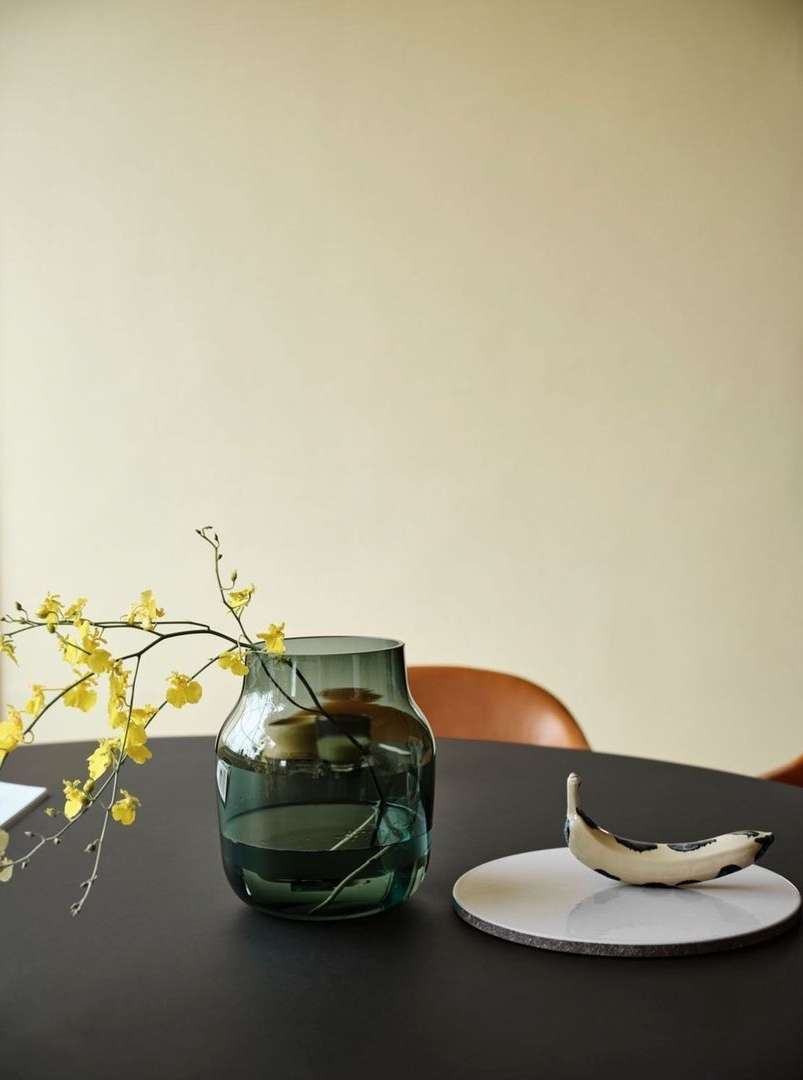 A vase with yellow flower stems sits on a black table, next to a ceramic dish, set against a creamy beige wall indoors.