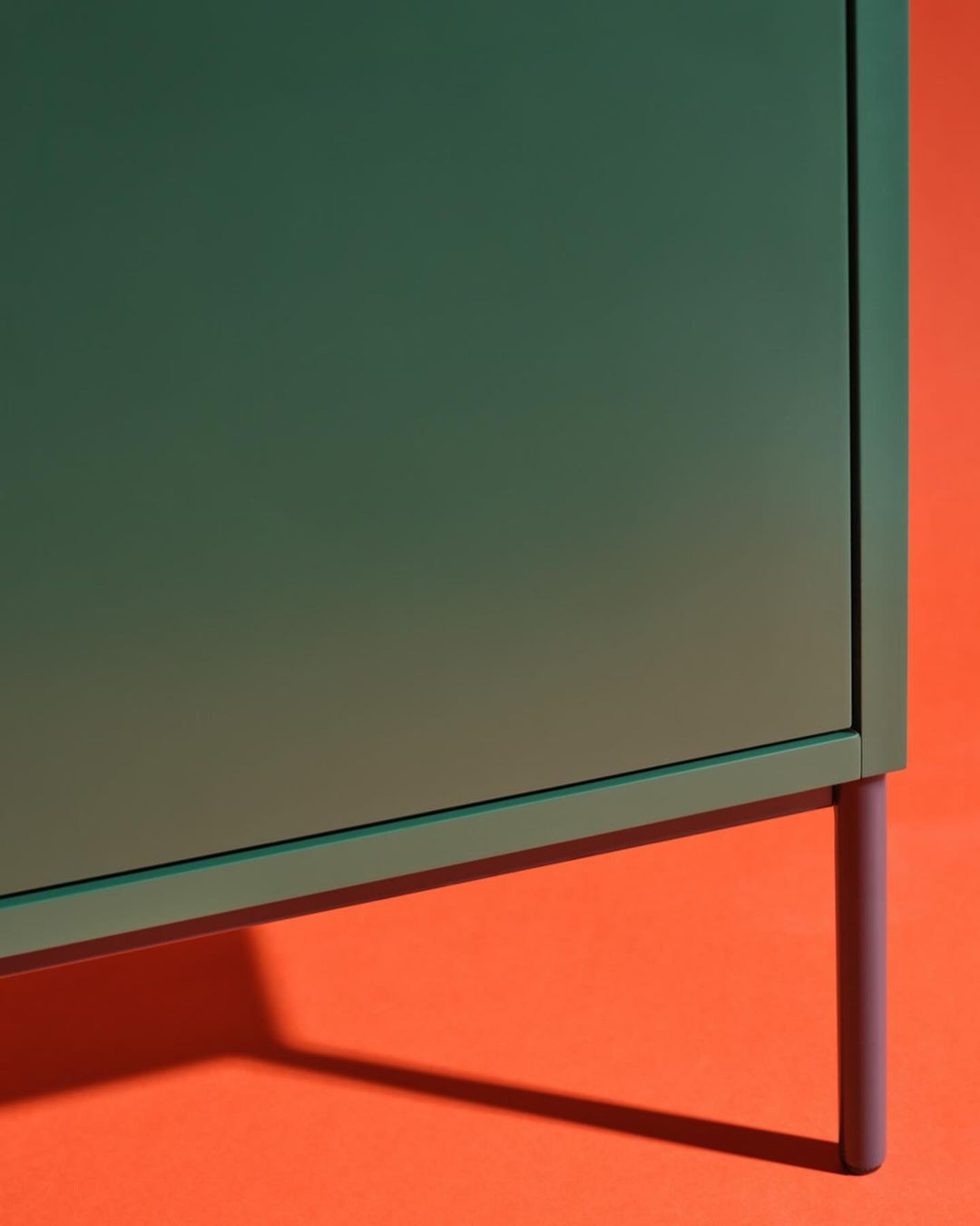 A green cabinet with a simple design stands on thin purple legs against an orange background, creating a minimalist and modern furniture display.