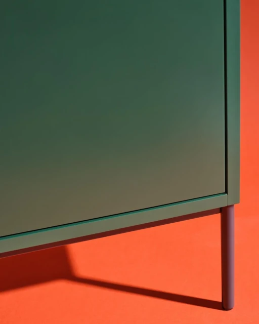 A green cabinet with a simple design stands on thin purple legs against an orange background, creating a minimalist and modern furniture display.