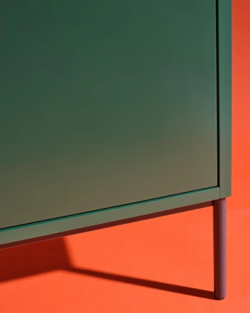 A green cabinet with a simple design stands on thin purple legs against an orange background, creating a minimalist and modern furniture display.